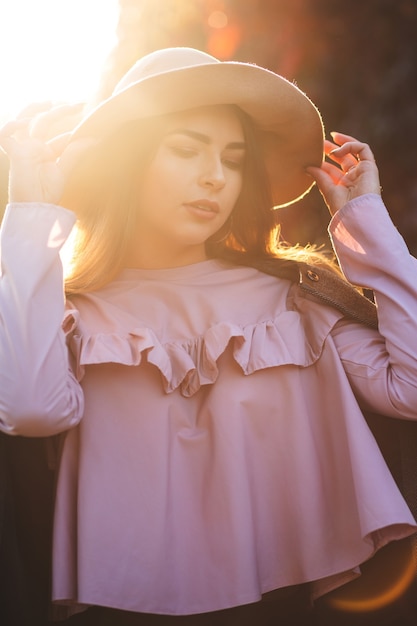 Elegante modelo bronceada con sombrero y blusa posando en los rayos del sol