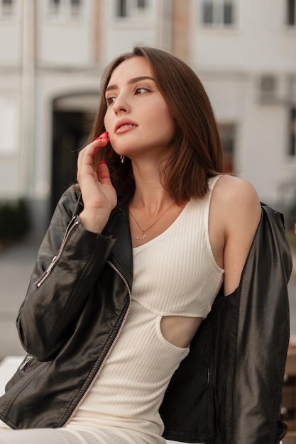Elegante modelo de jovem glamourosa com cabelo curto em uma elegante jaqueta de couro preta e vestido branco senta e relaxa na cidade
