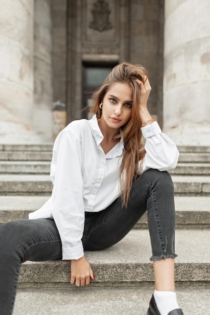 Elegante modelo de moda jovem linda em streetwear elegante com uma camisa branca e jeans preto senta-se nos degraus da cidade perto de edifícios antigos com colunas