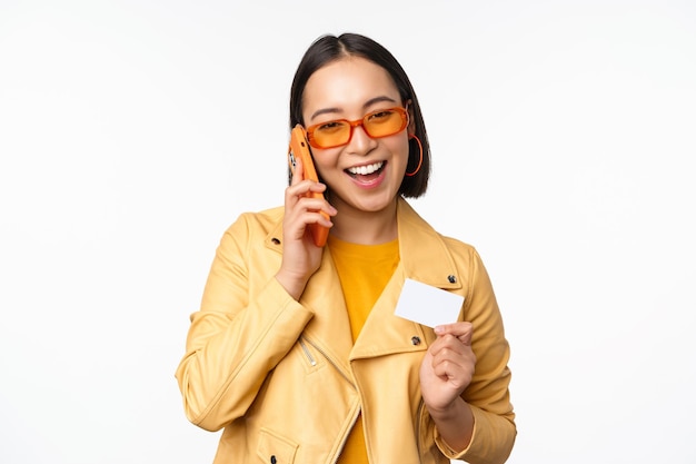 Elegante modelo femenino asiático hablando por teléfono inteligente y mostrando la tarjeta de crédito de pie sobre fondo blanco.