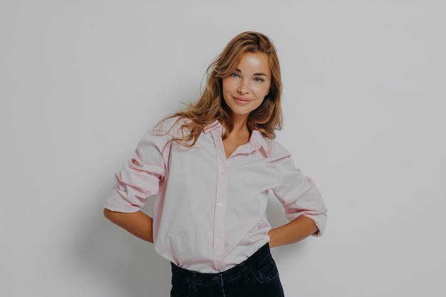 Elegante modelo femenino atractivo en camisa casual y jeans de pie con las manos cruzadas detrás de la espalda