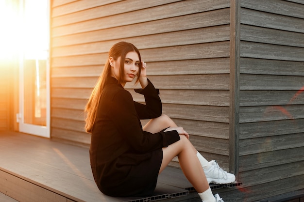 Elegante modelo de mujer joven y bonita en abrigo negro con elegantes zapatillas blancas se sienta cerca de una casa de madera al atardecer