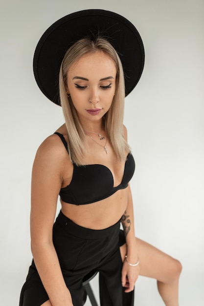 Elegante modelo de mujer joven con estilo con sombrero negro en lencería negra de moda con sujetador y pantalones se sienta en una silla en el estudio