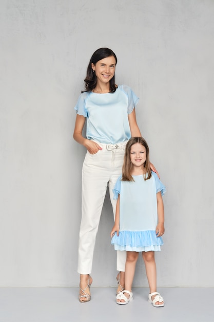 Elegante y moderna madre y su pequeña hija posando juntos cerca de la pared gris