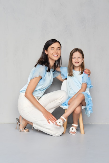 Elegante y moderna madre y su pequeña hija posando juntos cerca de la pared gris