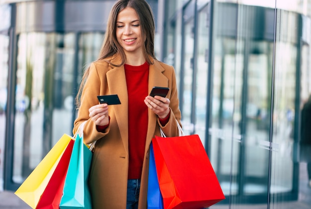 Elegante y moderna mujer joven hermosa con el uso de teléfonos inteligentes para la banca en línea y bolsas de compras mientras camina al aire libre después del centro comercial