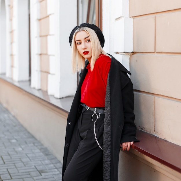 Elegante moderne junge Frau in einer Baskenmütze in einem roten Hemd mit einem modischen langen Mantel in der stilvollen Hose nahe einem Vintagen Gebäude am Herbsttag