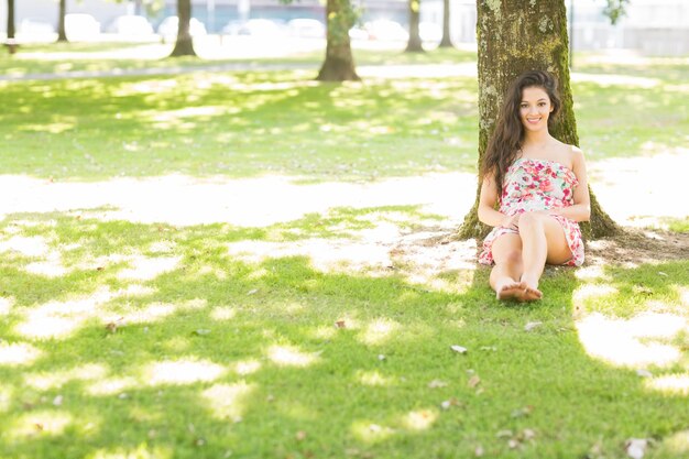 Foto elegante morena alegre sentada e encostada à árvore