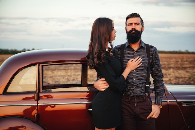 Una elegante morena barbuda con una camisa y pantalones con tirantes abraza a una joven esbelta con el pelo largo con un vestido corto negro cerca de un coche retro marrón en el fondo del campo