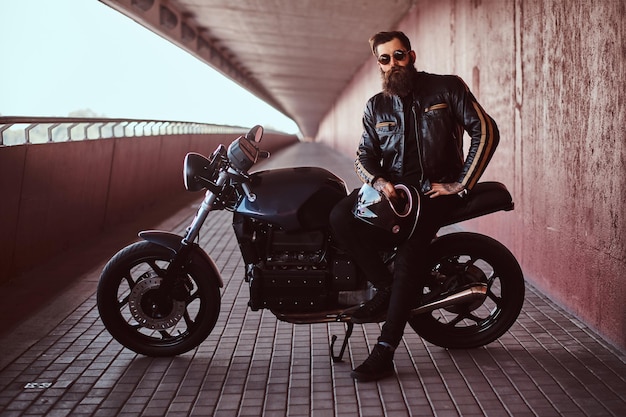 Elegante motociclista de moda vestido con una chaqueta de cuero negro con gafas de sol sostiene un casco sentado en su motocicleta retro hecha a medida, mirando una cámara.