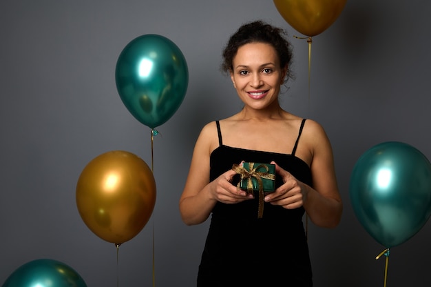 Elegante mujer africana en traje de noche sostiene un regalo de Navidad envuelto en papel verde brillante y cinta dorada, sonríe tiernamente con una gran sonrisa mirando a la cámara, contra globos de aire y fondo gris