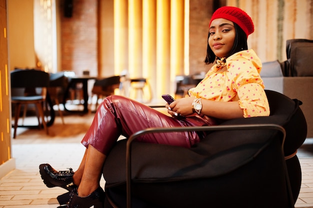 Elegante mujer afroamericana en boina francesa roja, blusa de lunares con cadena de cuello dorado grande y pantalones de cuero sentado en un puf con teléfono móvil.