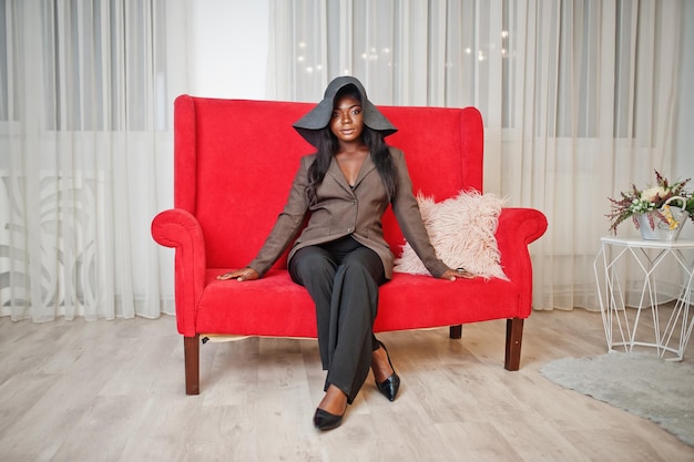 Elegante mujer afroamericana con ropa formal y sombrero sentada en un sofá rojo en la habitación blanca