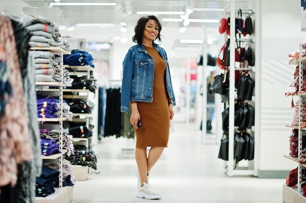 Elegante mujer afroamericana con vestido de túnica marrón y chaqueta de jeans posada en la tienda de ropa Es hora de ir de compras