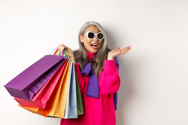 Elegante mujer asiática senior en gafas de sol yendo de compras en venta navideña, sosteniendo bolsas de papel y sonriendo, de pie sobre fondo blanco.