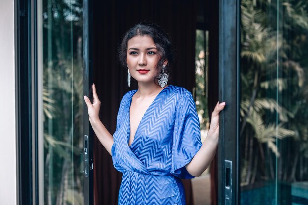 Elegante mujer asiática con vestido azul de cuello bajo abre puertas de vidrio en su villa; concepto de lujo exótico.