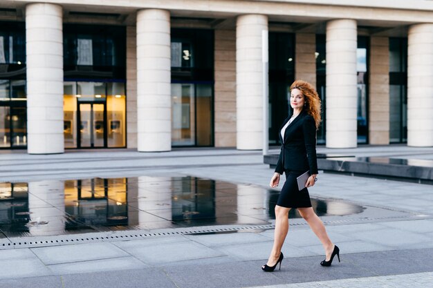 Elegante mujer delgada con traje negro y zapatos de tacón alto, con tableta en las manos yendo a trabajar