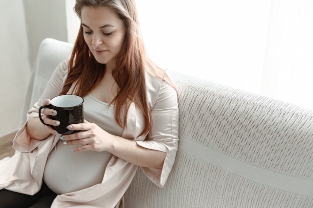 Elegante mujer embarazada con una gran barriga se sienta en el sofá en casa y bebe té o café.