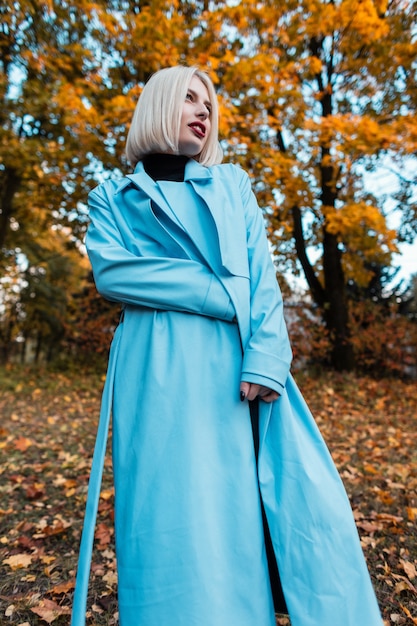 Elegante mujer hermosa con el pelo corto y rubio en un abrigo azul de moda posa en un bosque de otoño sobre un fondo de follaje de otoño amarillo