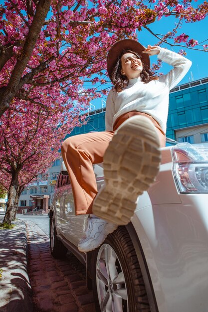 Elegante mujer hermosa sonriente de pie cerca del árbol de sakura floreciente del coche en el fondo se acerca la primavera