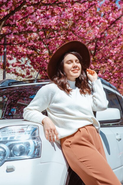 Elegante mujer hermosa sonriente de pie cerca del árbol de sakura floreciente del coche en el fondo se acerca la primavera