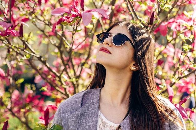 Elegante mujer hipster disfrutando del sol en flores de color rosa magnolia en un parque soleado niña con gafas de sol sonríe en el jardín botánico en el espacio de primavera para un momento alegre de texto