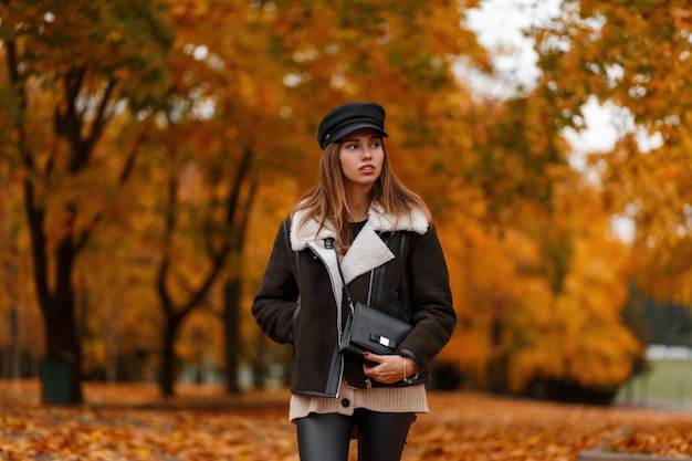 Elegante mujer joven y bonita con un elegante sombrero en una elegante chaqueta con una bolsa en el bosque sobre un fondo de hojas doradas. Linda chica camina en el parque. Colección de otoño de ropa de mujer de moda.