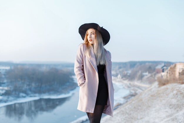 Elegante mujer joven con estilo en un elegante abrigo rosa con un elegante sombrero con un vestido de punto negro está de pie en una montaña en el río. Chica elegante disfruta del paisaje invernal.