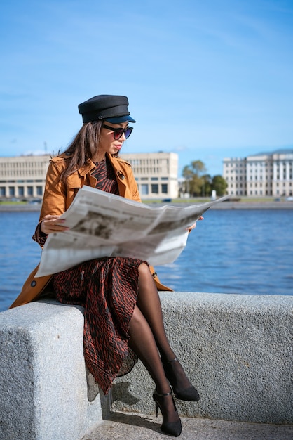 Elegante mujer joven de etnia caucásica lee periódicos frescos con gafas de sol y gorra negra beautif ...