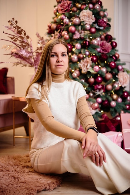 Una elegante mujer joven feliz con un traje de punto blanco de moda está sentada en una alfombra suave en una habitación acogedora cerca de un árbol de Navidad La modelo alegre de la chica de moda descansa en el estudio de Navidad