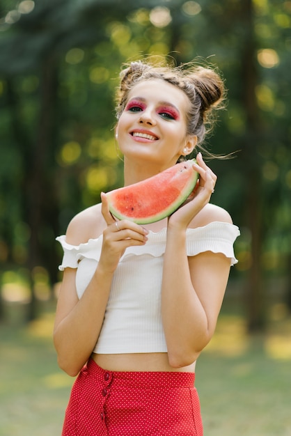 Elegante mujer joven hermosa con maquillaje jugoso brillante en forma de sandía sostiene un trozo de sandía en sus manos