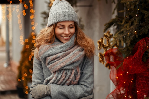 Elegante mujer joven de moda con una sonrisa feliz en ropa de punto con un sombrero gris y bufanda de moda en la calle en vacaciones de invierno cerca de las luces