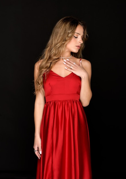 Elegante mujer joven preparada modelando un elegante vestido rojo de pie con la mano en el pecho y la cabeza apartada con los ojos cerrados sobre un fondo negro de estudio