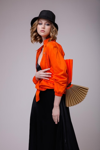 elegante mujer joven en un vestido negro sombrero camisa naranja bolso posando sobre fondo blanco