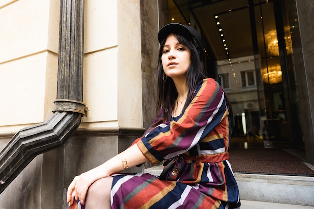 Elegante mujer joven con un vestido de rayas multicolores y un sombrero negro sentado en los escalones del hotel