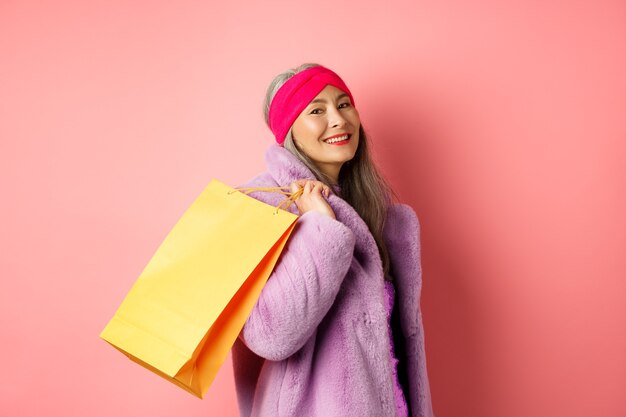 Elegante mujer mayor asiática que va de compras, lleva una bolsa de papel en el hombro y sonríe feliz a la cámara, de pie sobre un fondo rosa.