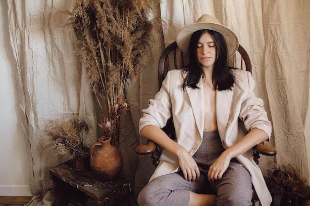 Elegante mujer de moda con traje y sombrero posando en una silla de madera en una habitación bohemia con hierba seca