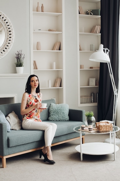 Elegante mujer mordiendo malvavisco en el sofá. Ropa casual y tacones sentados en el moderno sofá con cojines y mordiendo deliciosos malvaviscos.