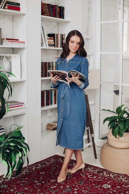 Elegante mujer morena hermosa en vestido de mezclilla y tacones leyendo una revista