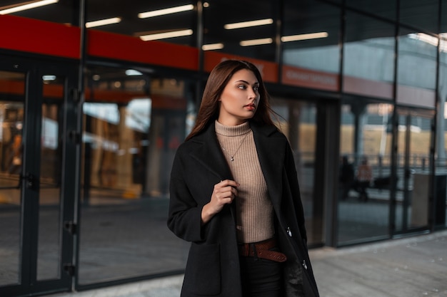 Elegante mujer morena joven y bonita con un abrigo negro de moda con una camisa beige de punto se encuentra cerca de un moderno edificio de cristal en la calle. Modelo de moda chica en ropa elegante en la ciudad.