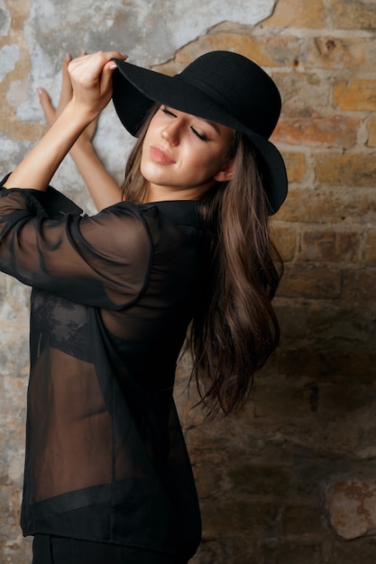 Elegante mujer morena con sombrero y blusa negra posando cerca de la pared del grunge