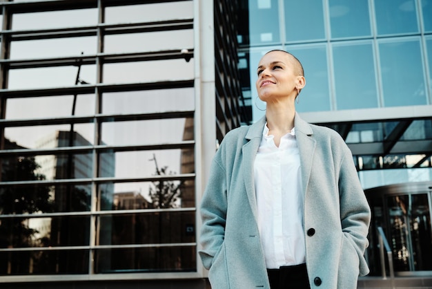 Foto elegante mujer de negocios mirando hacia otro lado y sonriendo mientras está de pie afuera en la calle con edificios de oficinas en el fondo. concepto de negocio.