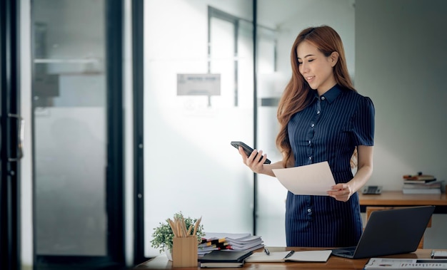 Elegante mujer de negocios de pie en la oficina con teléfono móvil y papeleo