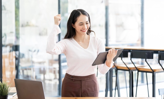 Elegante mujer de negocios sentada en la oficina con tableta digital Emocionada mujer de negocios asiática levantando la mano para felicitar mientras trabaja en una computadora portátil en la oficina