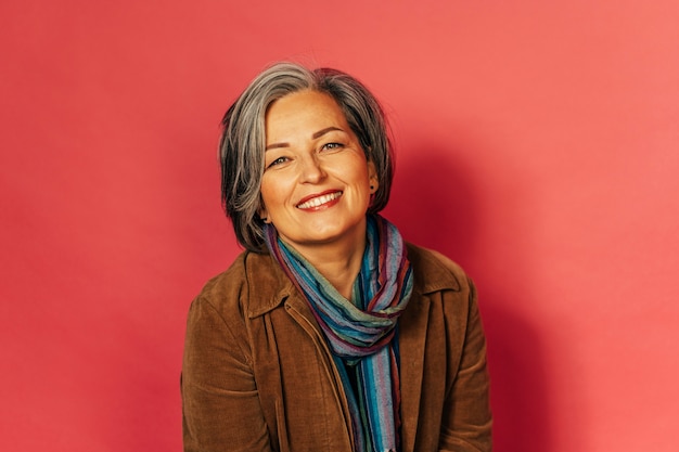 Elegante mujer de pelo gris sonríe ampliamente posando en estudio en pared rosa. Encantadora bella dama madura mira a la cámara. Concepto de glamour. Imagen teñida