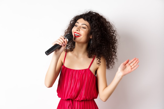 Elegante mujer de pelo rizado en vestido rojo cantando en el micrófono, mirando a un lado y sonriendo feliz, de pie sobre fondo blanco.