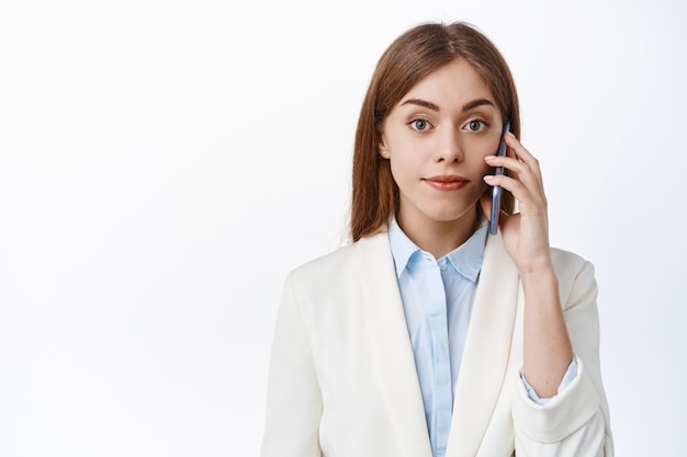 Elegante mujer profesional habla por teléfono móvil, mira al frente seguro y decidido, haciendo una llamada en el trabajo, de pie sobre una pared blanca