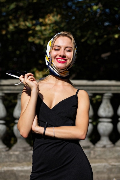 Elegante mujer rubia sonriente con labios rojos con mantón y vestido negro posando con la luz del sol