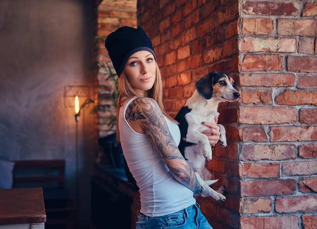 Una elegante mujer rubia tatuada con camiseta y jeans sostiene un lindo perro.