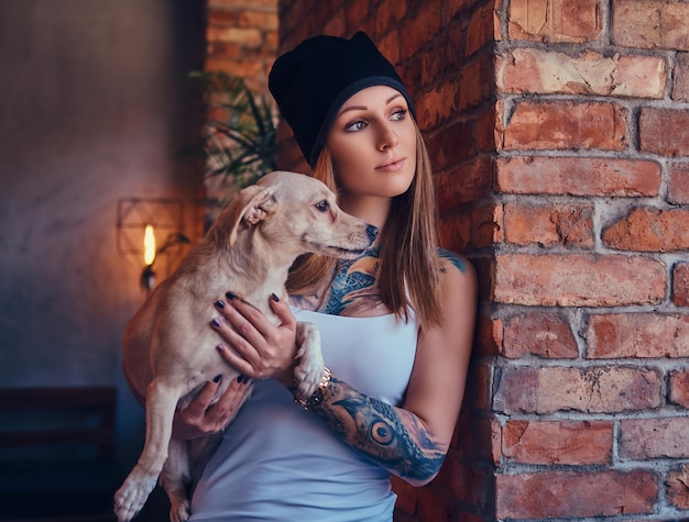 Una elegante mujer rubia tatuada con camiseta y jeans sostiene un lindo perro.